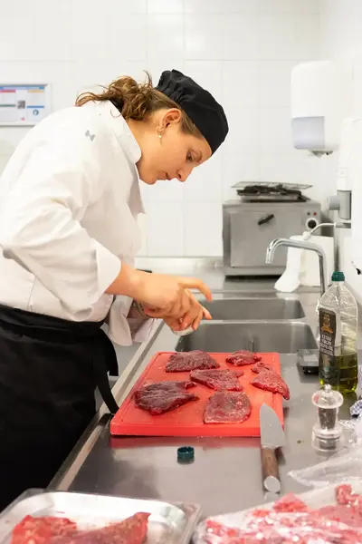 Kok in witte jas en zwarte muts snijdt rood vlees op een rode snijplank in een professionele keuken.