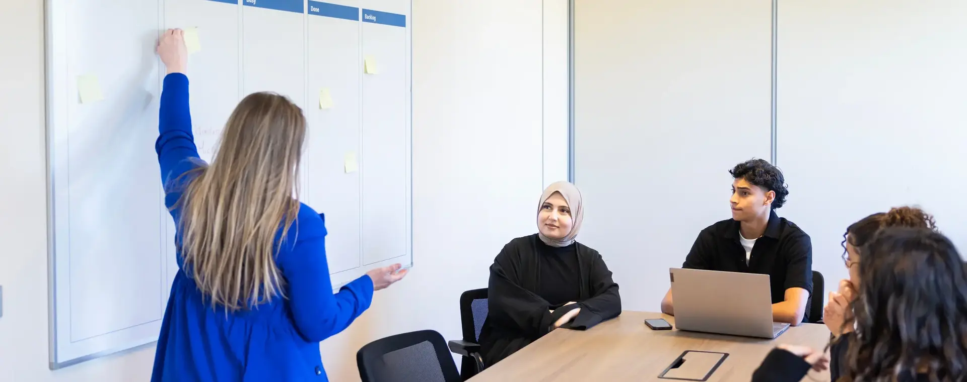 Studente wijst iets aan op een whiteboard en drie mensen zitten aan een vergadertafel.