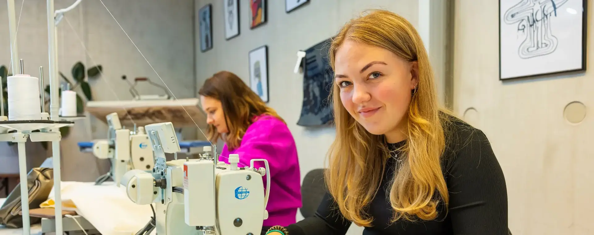 Twee vrouwen zitten achter naaimachines te werken in een hippe werkplaats of klaslokaal. De vrouw op de voorgrond lacht in de camera terwijl ze bezig is.