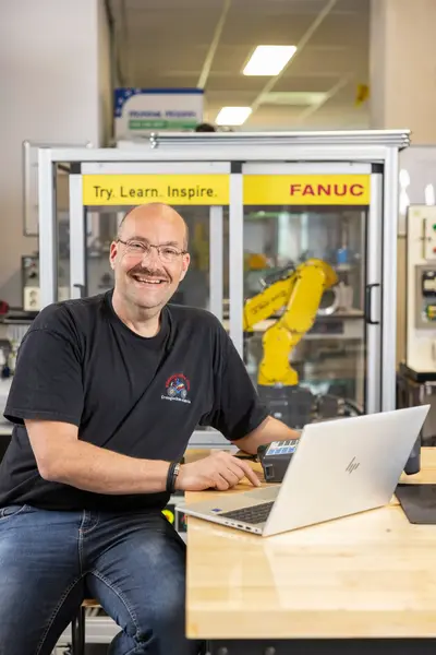 Man in T-shirt achter laptop in fabriekssetting met robotarm