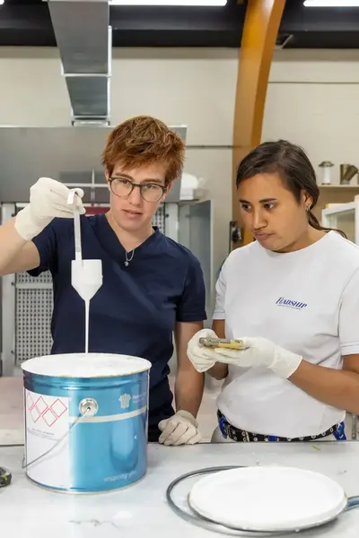 Twee studenten en een docent staan bij een werktafel met een emmer verf en kijken hoe een student met een speciale lepel verf uit de emmer haalt. 