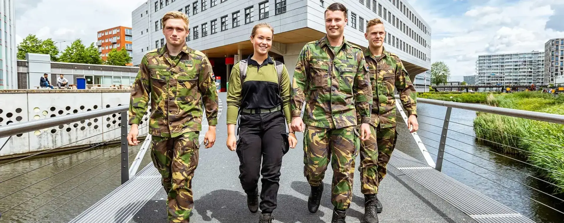 Vier studenten in militaire uniformen lopen lachend over een brug bij MBO College Zuidoost.