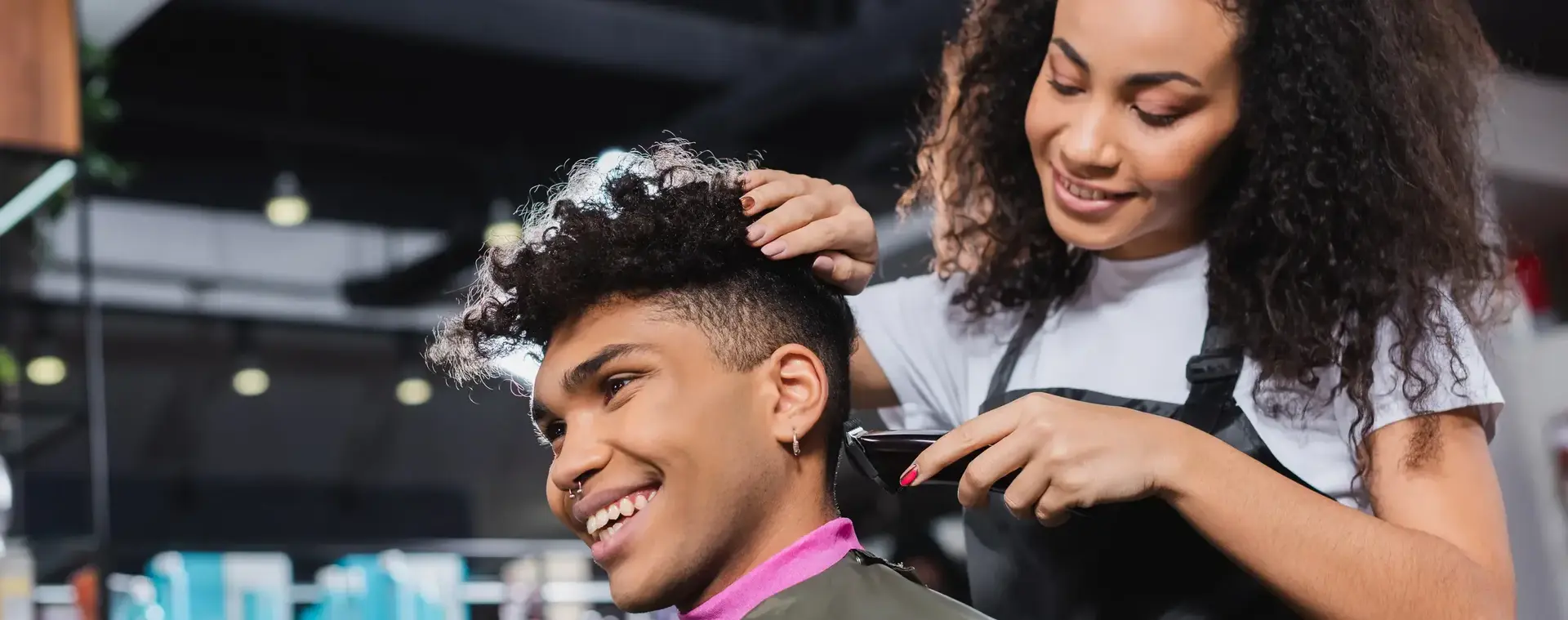 Student knipt met een tondeuse het haar van een jongen in een kapsalon. Ze lacht terwijl ze geconcentreerd werkt aan het kapsel van haar klant.