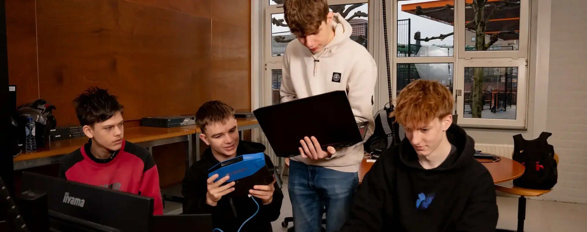 Vier studenten werken samen in een klaslokaal aan laptops en IT-apparatuur, terwijl één student een netwerkapparaat bekijkt.