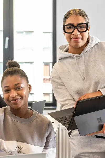 Drie studenten werken in een klaslokaal, twee studenten kijken lachend naar de camera en één student werkt geconcentreerd op zijn laptop.