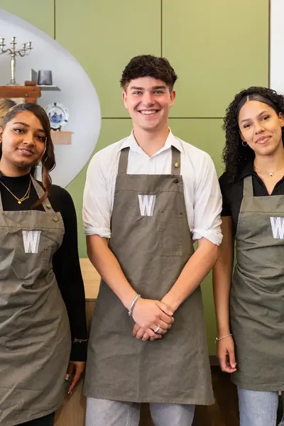 Drie medewerkers staan in een horecazaak en dragen een grijs schort met een witte "W" erop.