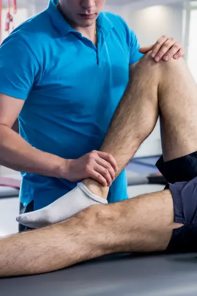 Een man ligt in een gymzaal en andere man beweegt zijn been in verband met een blessure.