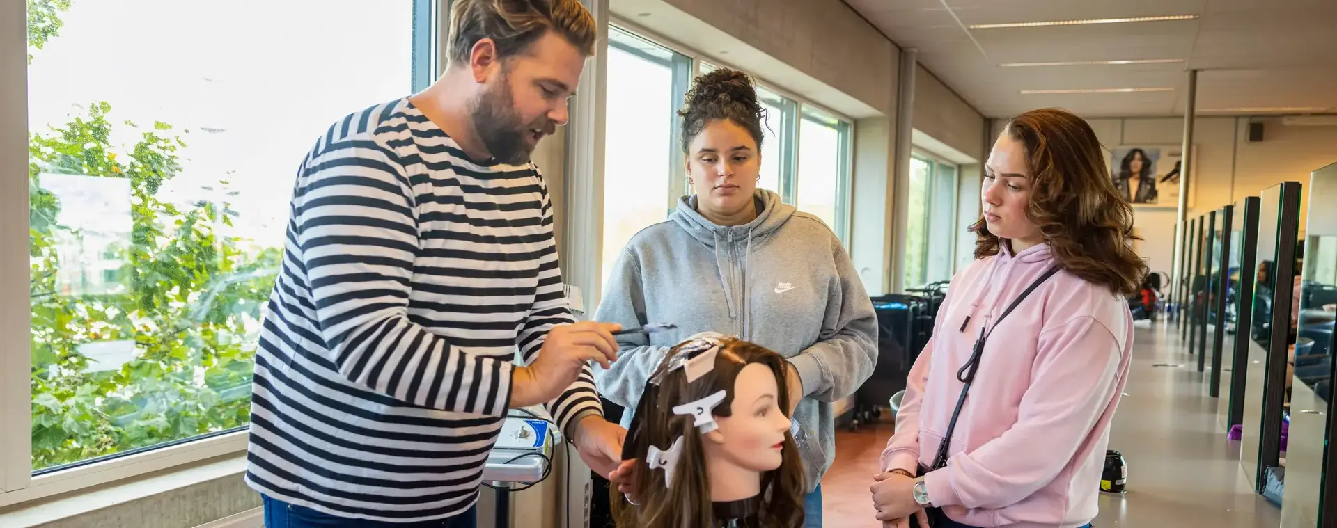 Docent geeft instructie over haartechnieken aan twee studenten die aandachtig meekijken, terwijl hij werkt aan een oefenhoofd in een kapsalonklaslokaal met grote ramen.
