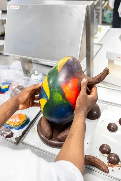 Een student werkt geconcentreerd aan een kleurrijk chocolade ei, omringd door vormen van gesmolten en gevormde chocolade.