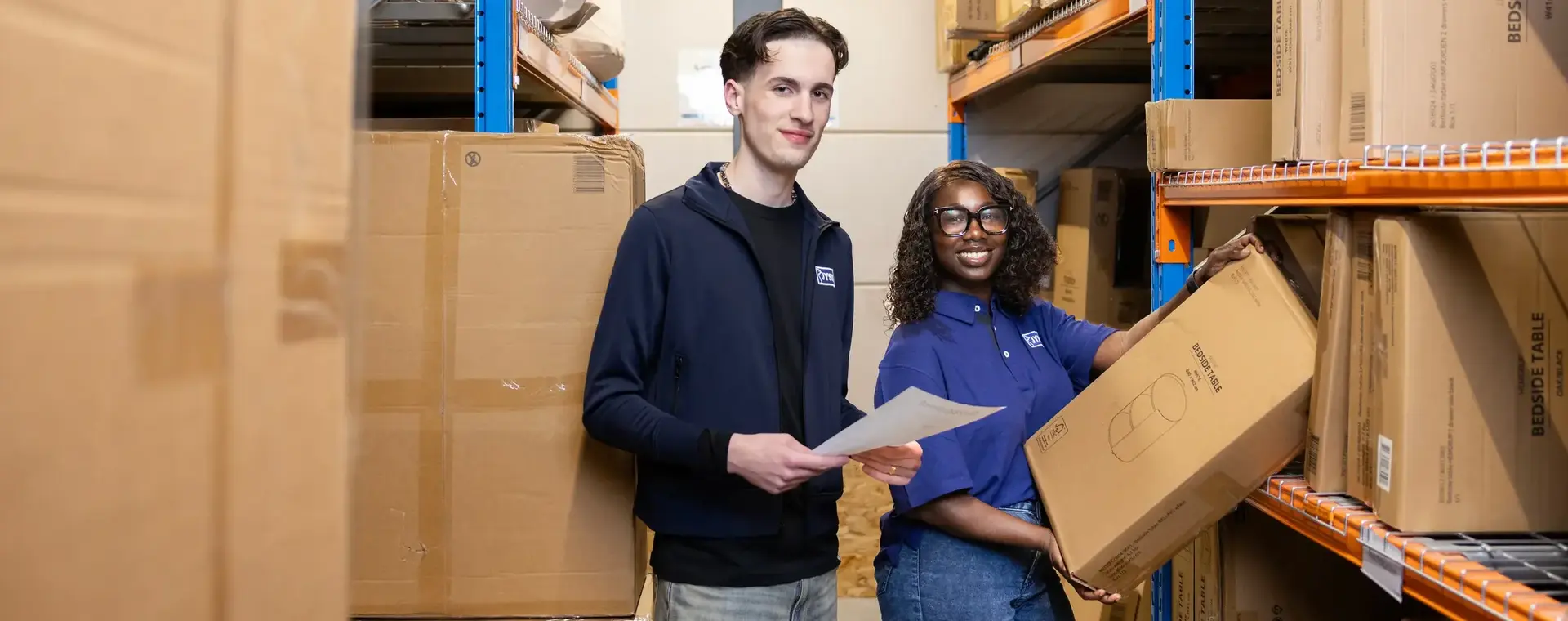 Twee studenten controleren samen de voorraad in een magazijn tussen dozen en stellingen
