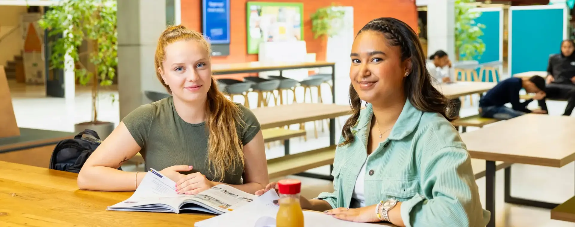 Twee studenten zitten in een open studieruimte aan een tafel met boeken en een flesje drinken.