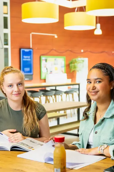 Twee studenten zitten in een open studieruimte aan een tafel met boeken en een flesje drinken.