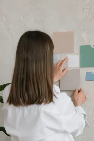 Student hangt kleurstalen op aan een muur tijdens een ontwerpoefening. Ze draagt een witte blouse en staat naast een kamerplant.
