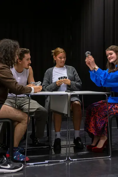 Vier studenten zitten aan een tafel op een podium en oefenen een toneelscène.