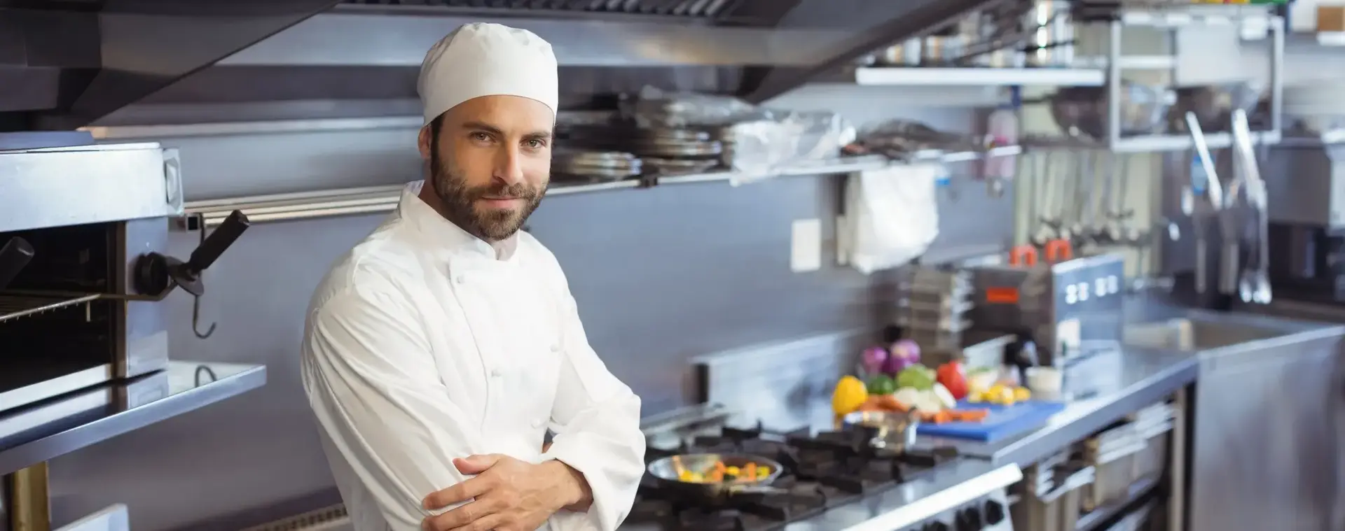 Een kok met koksmuts staat in een grote keuken van een restaurant. Hij heeft zijn armen over elkaar geslagen en kijkt glimlachend de camera in.