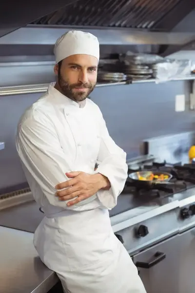 Een kok met koksmuts staat in een grote keuken van een restaurant. Hij heeft zijn armen over elkaar geslagen en kijkt glimlachend de camera in.