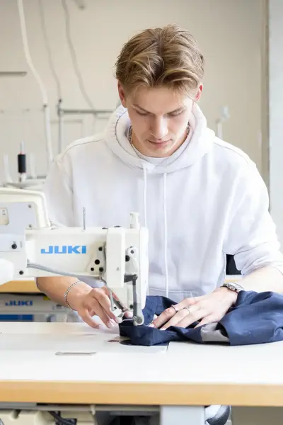 Student werkt geconcentreerd achter een naaimachine in het atelier en stikt donkerblauwe stof. Op tafel liggen schaar en materialen voor het ontwerp.