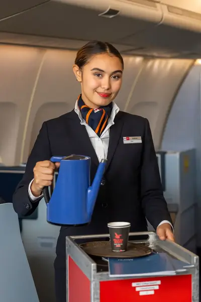 Vriendelijke stewardess schenkt drankjes in tijdens vlucht en staat met een trolley in het gangpad van het vliegtuig.