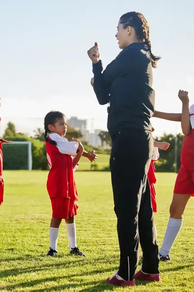 Vrouwelijke voetbaltrainer doet met vier kinderen in voetbaltenues stretch oefeningen op het voetbalveld om op te warmen.