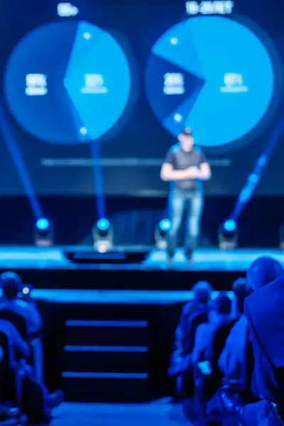 Man houdt presentatie op podium in een grote zaal met mensen.