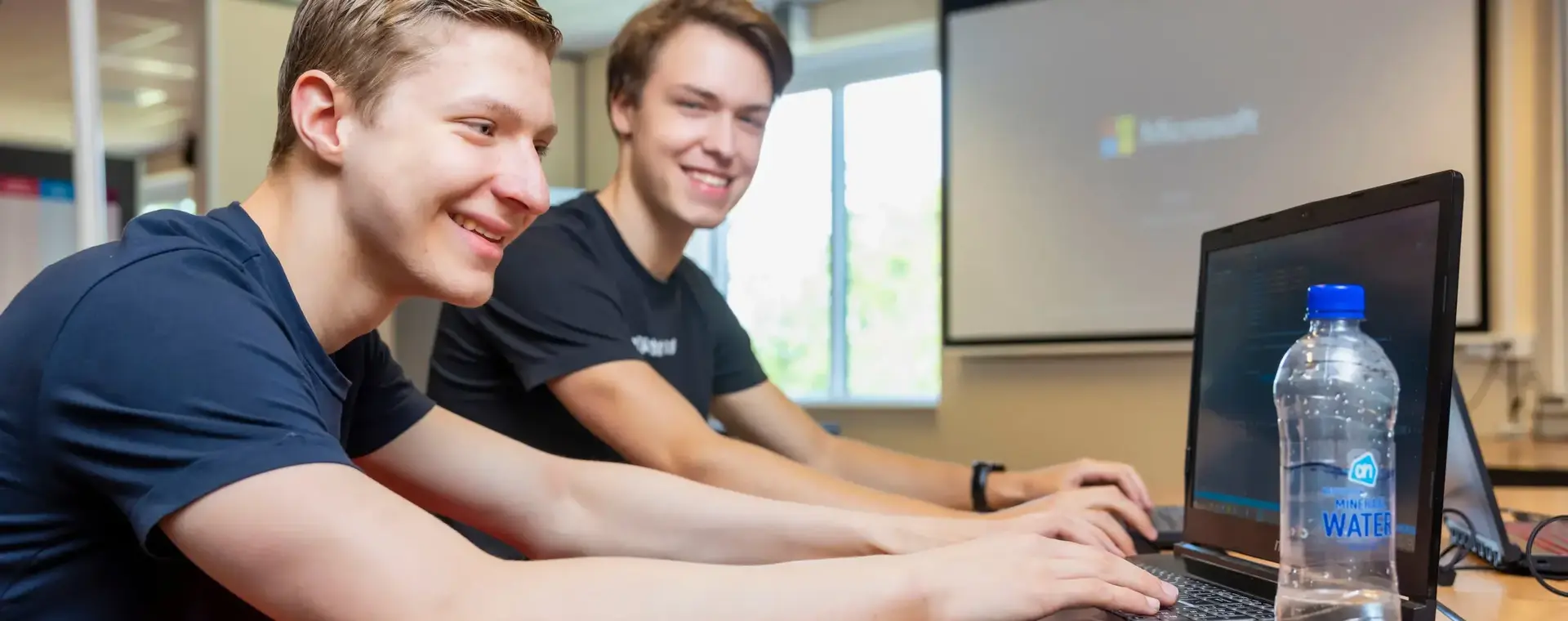 Twee studenten werken enthousiast samen achter een laptop in een klaslokaal, met een beamer op de achtergrond.