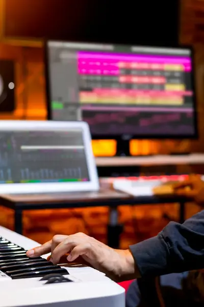Student produceert muziek in een opnamestudio, werkend aan een toetsenbord met muzieksoftware zichtbaar op twee schermen.