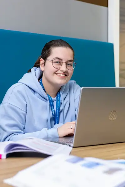 Student zit lachend achter een laptop in de studieruimte en werkt samen met een klasgenoot. Op tafel liggen open boeken en schoolspullen.