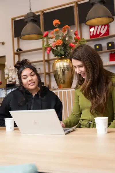 Twee vrouwen zitten samen aan een houten tafel in een koffiebar, lachend terwijl ze naar een laptop kijken. Op de achtergrond staan een bloemenvaas.