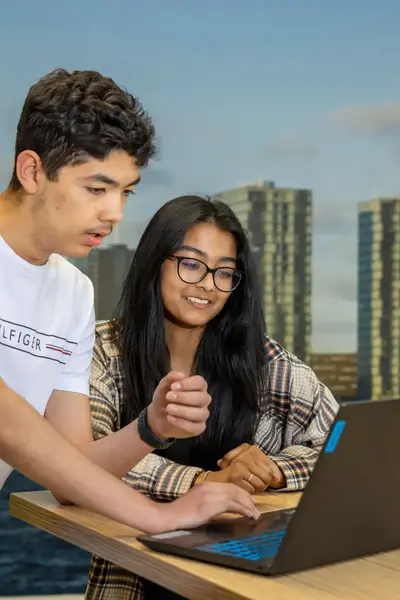 Twee studenten werken samen achter een laptop, waarbij één student iets uitlegt terwijl de ander glimlachend toekijkt, met een skyline van hoge gebouwen en water op de achtergrond.