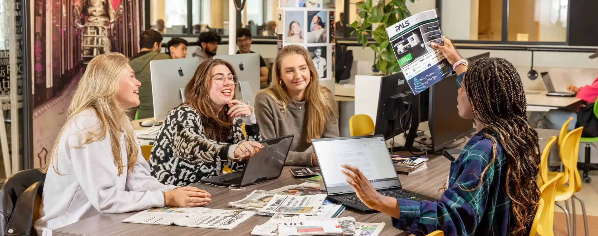 Er zitten vier studenten aan een vergadertafel. Er liggen allemaal magazines op de tafel en er staan een laptop open. Ze zijn de magazines aan het bespreken.