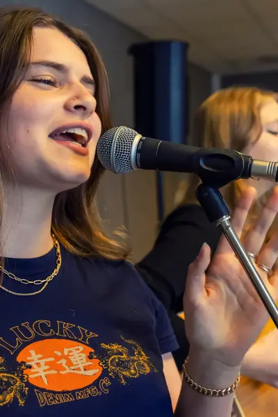 Student zingt in een microfoon tijdens een muziekles, met een medestudent op gitaar op de achtergrond.