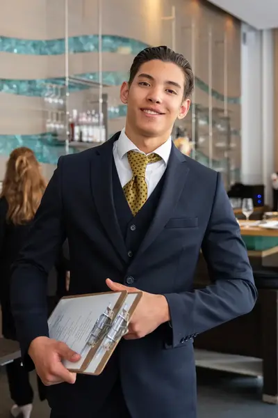 Jonge hotelmedewerker in donker pak en stropdas houdt een notitieblok vast en glimlacht. In de hotelruimte werken collega’s.