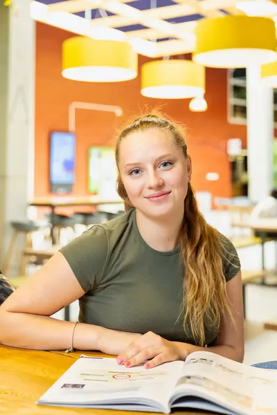 Student met lang blond haar zit in een studieruimte en kijkt glimlachend op.