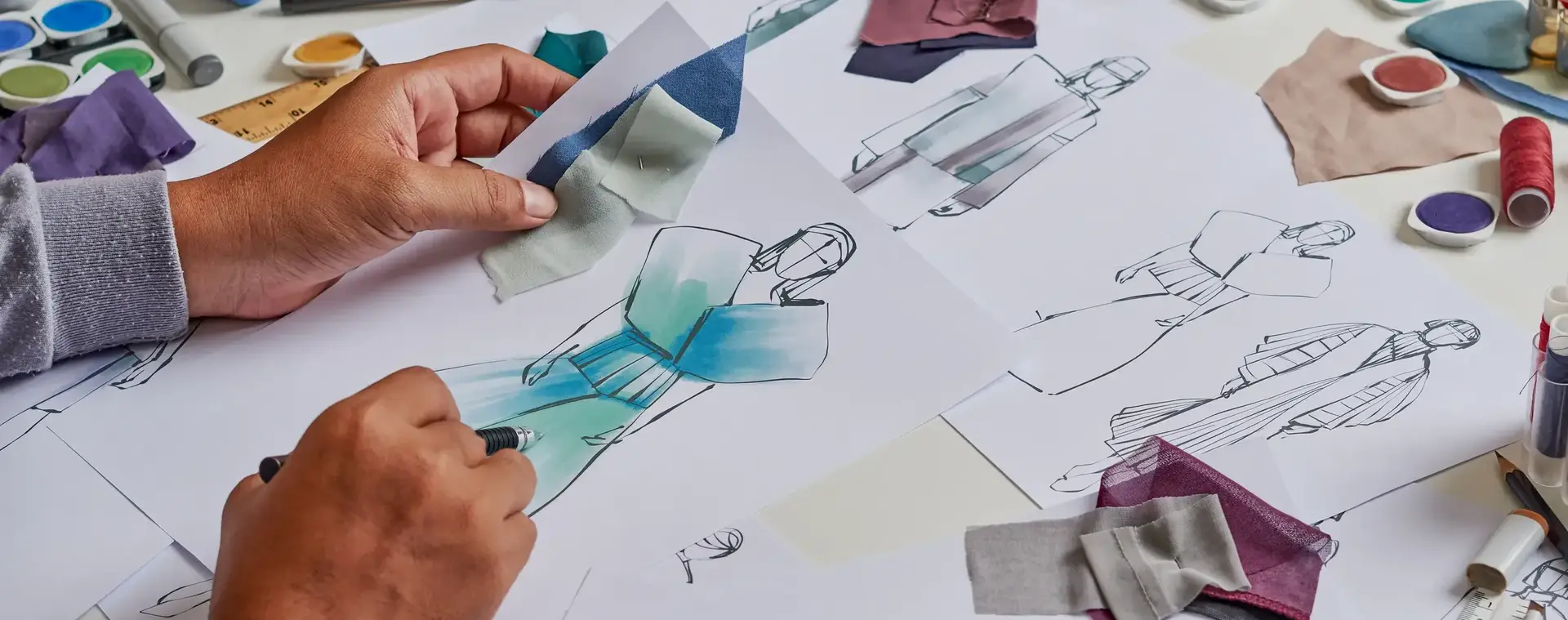 Iemand is mode aan het ontwerpen. Je ziet twee handen met een potlood die een jurk aan het designen is. Op de tafel liggen meerdere designs.