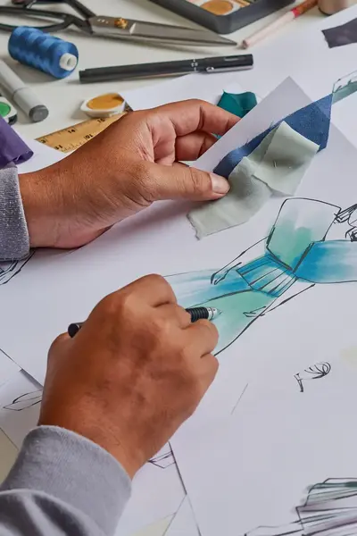 Iemand is mode aan het ontwerpen. Je ziet twee handen met een potlood die een jurk aan het designen is. Op de tafel liggen meerdere designs.