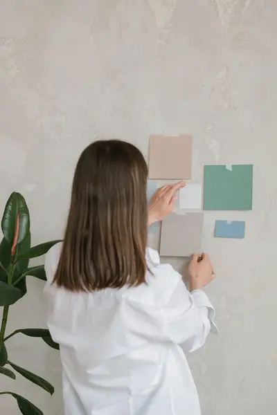 Student hangt kleurstalen op aan een muur tijdens een ontwerpoefening. Ze draagt een witte blouse en staat naast een groene kamerplant.