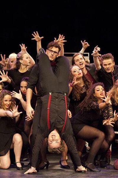 Studenten voeren een expressieve theatervoorstelling op, waarbij één student ondersteboven wordt vastgehouden door een medestudent, omringd door klasgenoten in theatrale poses.
