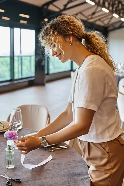 Een jonge vrouw is bezig met het decoreren van een gedekte tafel in een sfeervolle ruimte met grote ramen. Ze plaatst een lint om een vaasje met bloemen.