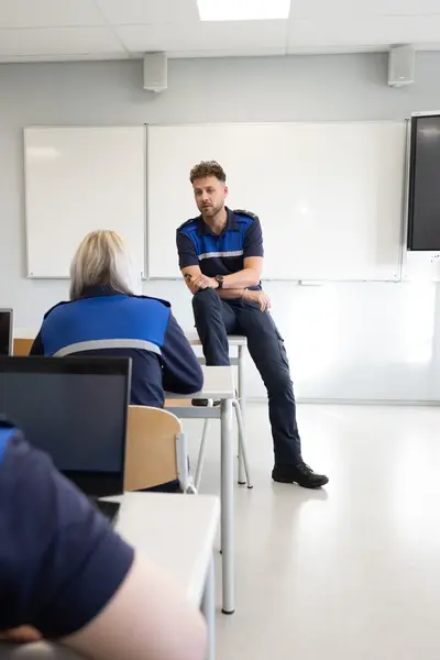 Docent veiligheidscollege zit nonchalant op de desk voor whiteboard in een klas met studenten in uniform