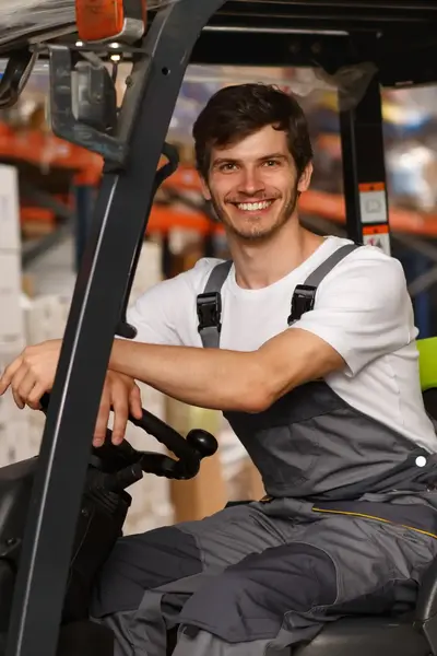 Magazijnmedewerker lacht terwijl hij op een heftruck zit tussen stapels dozen in een magazijn.