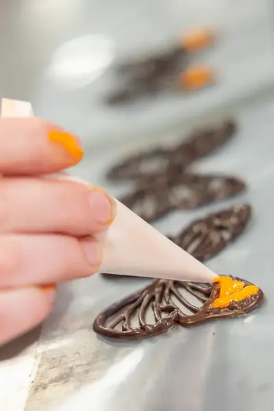 Close-up van een hand die met precisie oranje details aanbrengt op een vlinder chocoladestuk met behulp van een spuitzakje.