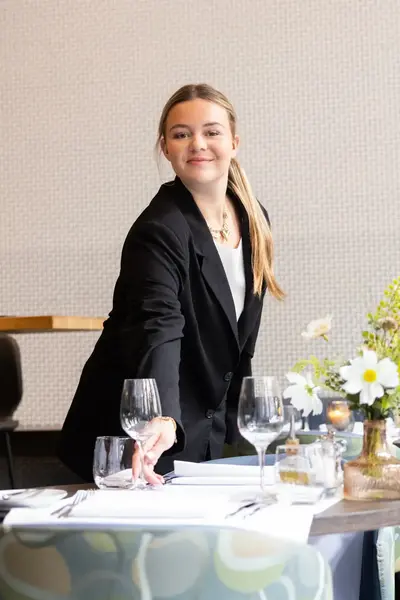 Medewerker in een zwarte blazer dekt een tafel in een restaurant. Op tafel staan glazen, servetten en bloemen in een vaas. Op de achtergrond staan barkrukken.