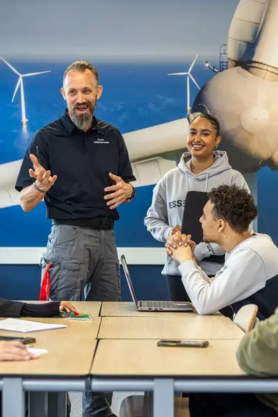 Docent geeft les aan een groep studenten in een lokaal met een grote afbeelding van windmolens op de muur. De studenten luisteren en lachen tijdens het gesprek. 