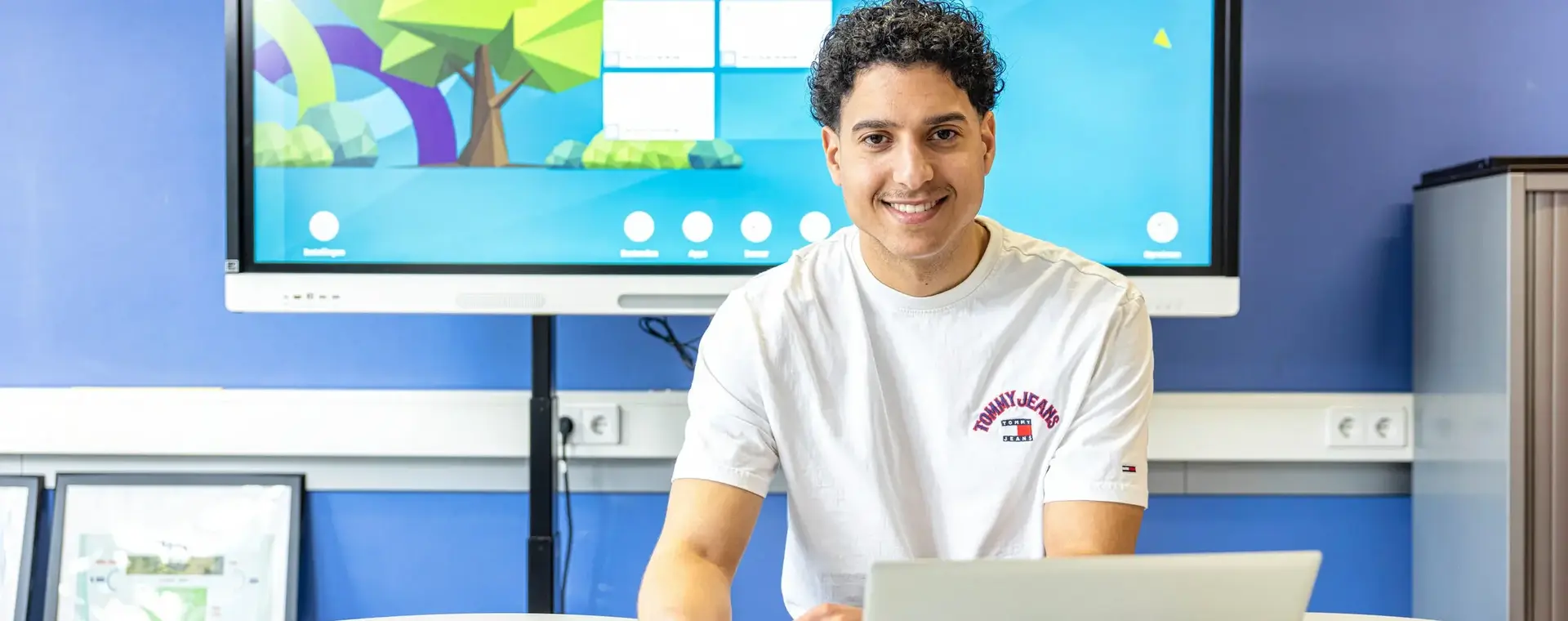 Docent zit aan een tafel met een laptop voor een digitaal schoolbord en kijkt lachend in de camera.