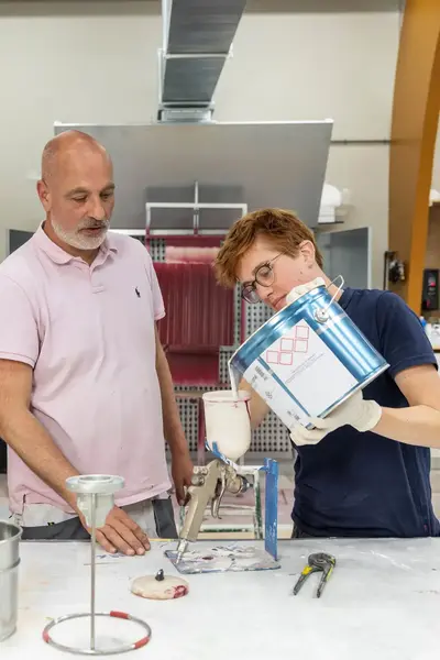 Twee studenten en docent staan bij een werktafel. Een student giet speciale verf uit een emmer in een onderdeel van het spuitapparaat om coating mee te kunnen aanbrengen.