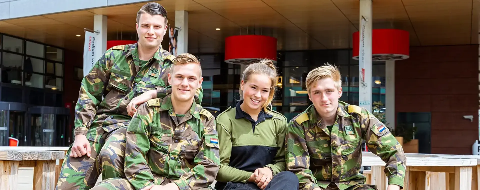 Vier studenten in legeruniform zitten en poseren lachend op houten banken buiten.