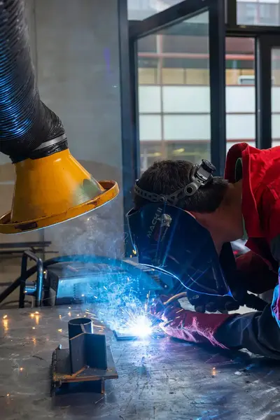 Student met lasmasker en veiligheidskleding staat gebogen te lassen