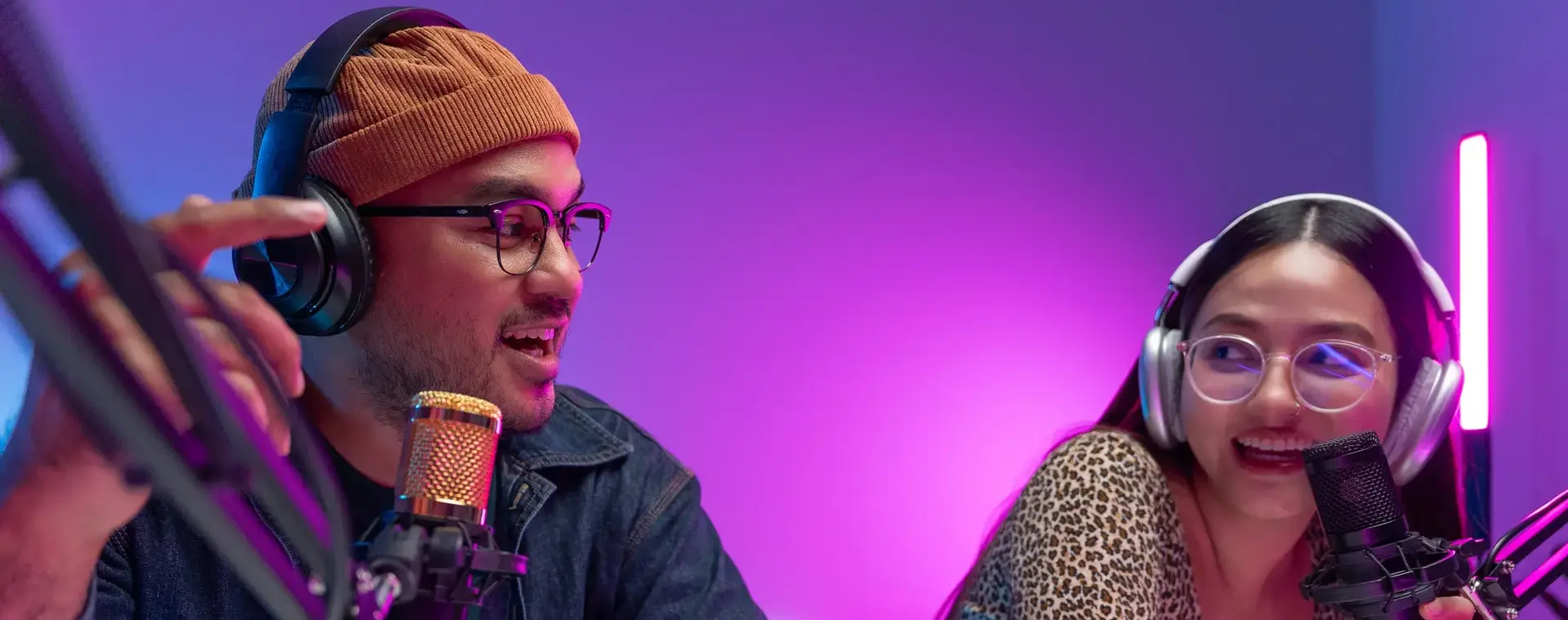 Een man en vrouw zitten in een radio studio achter twee microfoons met elkaar te praten en radio te maken.