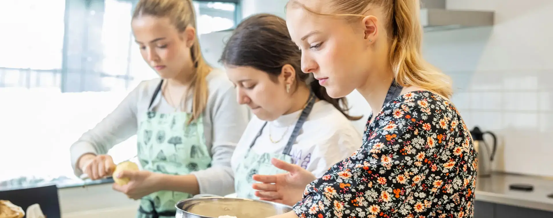 Drie jonge vrouwen bereiden samen eten in een keuken. Ze dragen schorten en zijn geconcentreerd bezig met het schillen van fruit en het mengen van ingrediënten.