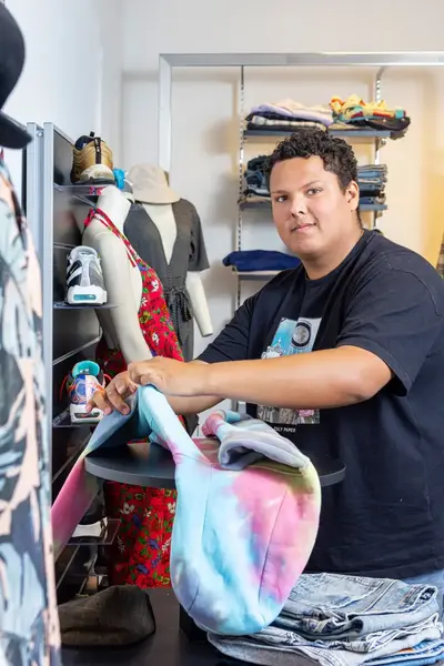 Student vouwt kleding in een modewinkel met rekken vol kleding, schoenen en paspoppen op de achtergrond.